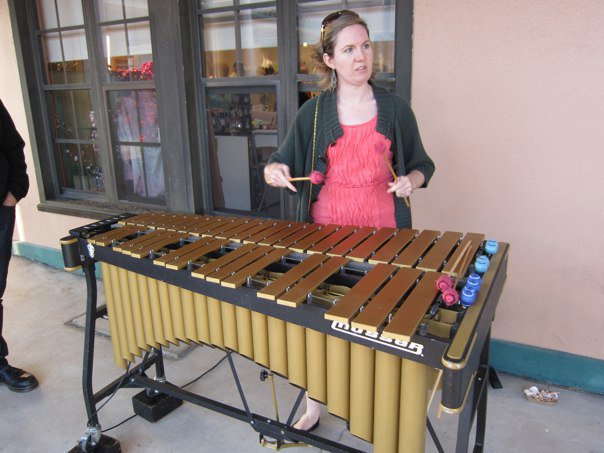 Rachel Moore, dressed in a pink top and green sweater, plays a gold vibraphone